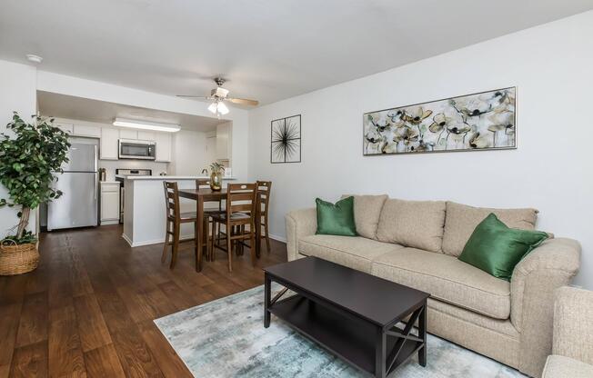 A cozy living room featuring a beige sofa with green accent pillows, a dark coffee table, and a light area rug. In the background, there is a dining area with a wooden table and chairs, and a kitchen with white cabinetry and a refrigerator. Natural light brightens the space, complemented by decorative wall art.