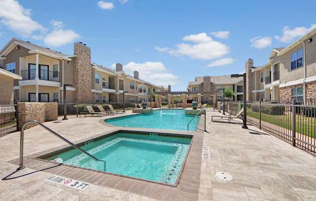 A spacious resort-style pool area here at Mission Green featuring a sparkling swimming pool and spa, expansive stone pool deck, metal fencing, comfortable lounge chairs, pergola accents, and surrounding apartment buildings beneath a bright blue sky, creating an inviting community space for relaxation and leisure.