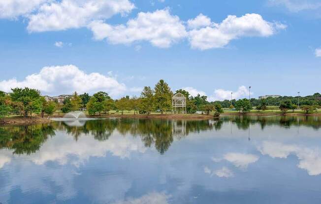 A serene lake with a fountain in the middle, surrounded by trees and a clear blue sky.