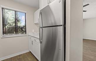 A stainless steel refrigerator in a kitchen with wooden flooring and a window with blinds.