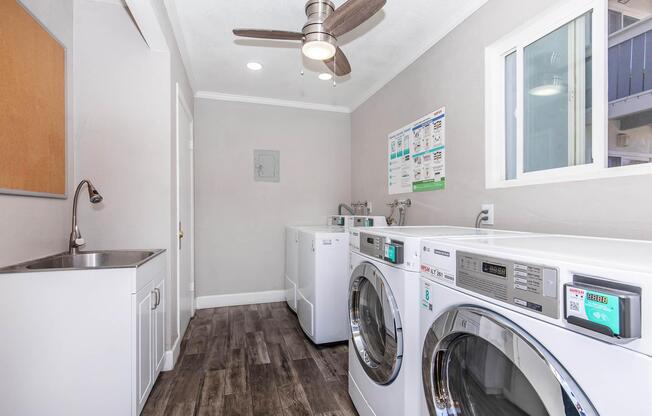 A clean and modern laundry room featuring multiple washing machines and dryers. There's a utility sink, a ceiling fan, and a window providing natural light. The walls are painted light gray, and there's a bulletin board with information on the wall. The floor is made of dark wood-like tiles.