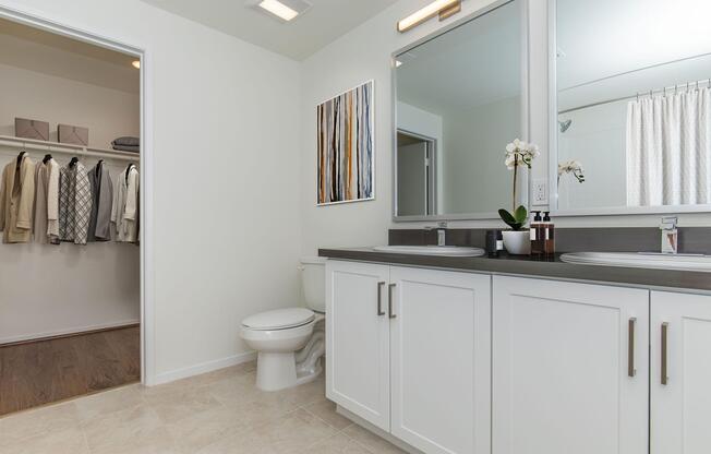 A clean, modern bathroom featuring two sinks with a dark countertop, a large mirror, and an orchid plant. The space is well-lit, with light-colored walls and tile flooring. To the side, a closet is visible, organized with neatly hung clothing items. A white shower curtain is seen in the background.