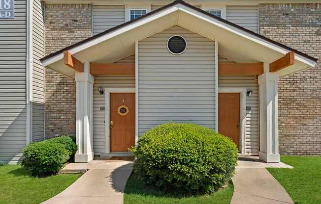 A welcoming exterior entryway here at Summertree Place featuring a covered gabled porch with neutral siding, brick accents, and two private front doors framed by neatly trimmed shrubs, creating a clean and inviting first impression along a well-maintained walkway.