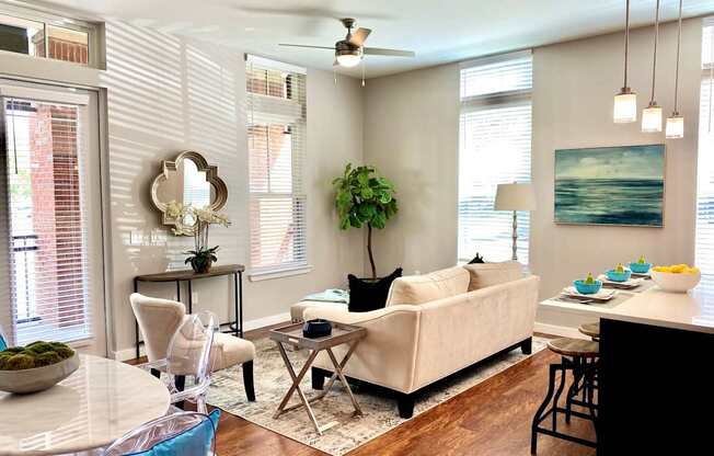 A living room with two windows, hardwood-style flooring, a ceiling fan, double doors leading to a private patio, and access to a dining area and kitchen.