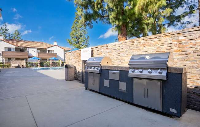 A BBQ grill is on a patio with a stone wall and trees in the background.