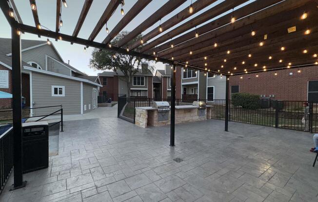 A patio area with a concrete floor and wooden beams overhead, adorned with string lights. In the center, there's a barbecue grill next to a stone structure. Surrounding the space are houses with various architectural styles and a fenced area. The setting suggests a cozy outdoor gathering space.