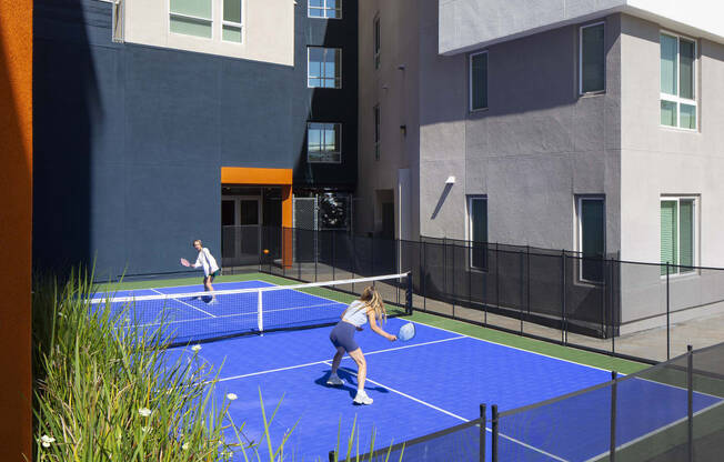 two women playing tennis on a blue court in front of a building