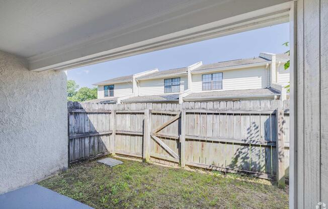 View from a patio or porch showing a fenced yard with grass and a small slab of concrete. In the background, residential buildings can be seen with windows and a roofline. The scene is bright and sunny, suggesting a warm day.