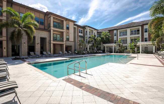 A swimming pool surrounded by a tiled patio and lounge chairs in front of a building with palm trees.