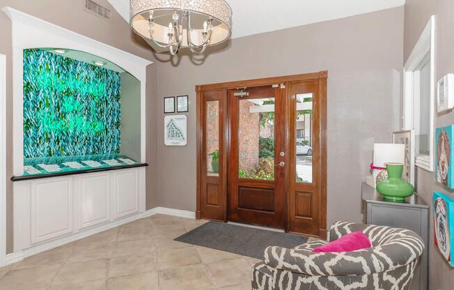 A well-decorated entryway featuring a stylish chandelier, a vibrant green art installation on the wall, and a seating area with a patterned chair. There's a wooden front door with glass panels, a tiled floor, and decorative accents like colorful artwork and a green lamp.