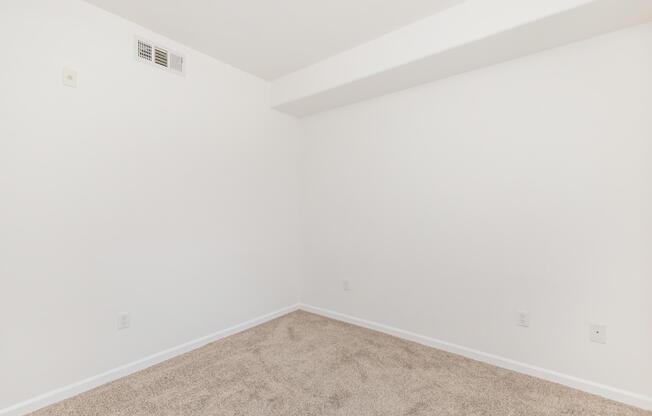 Empty room with light-colored walls and a carpeted floor. There are no furniture items or decor, creating a spacious and minimalist appearance. A small air vent is visible on the upper left wall. Natural light is not present in the image.