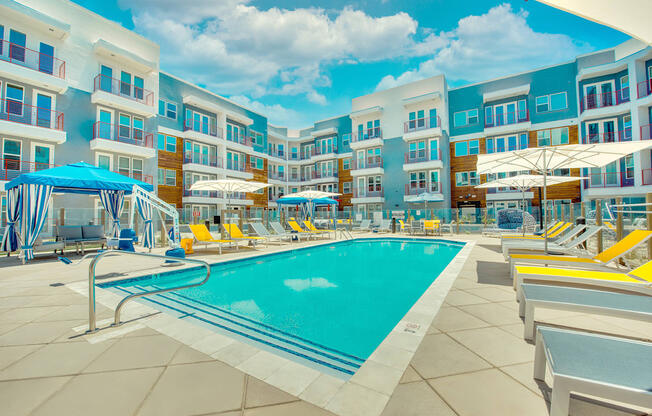 A swimming pool surrounded by sun loungers and umbrellas in front of a multi-story hotel.