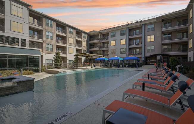 A swimming pool surrounded by sun loungers and umbrellas in front of apartment buildings.