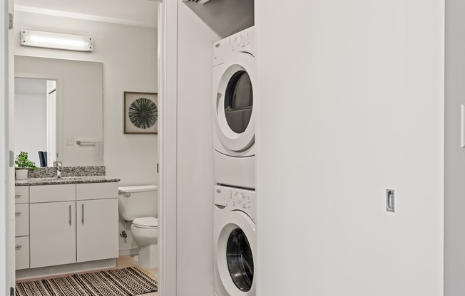 A white washing machine is in a laundry room with a toilet and sink in the background.