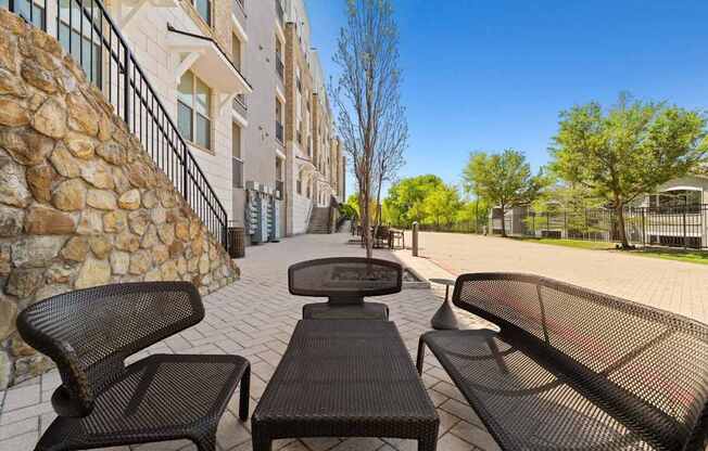A stone wall separates a patio with three black chairs from a building.