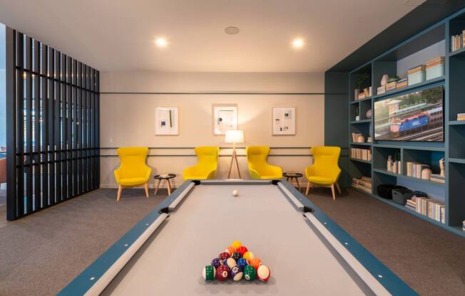 A pool table with a yellow chair and a blue bookshelf.