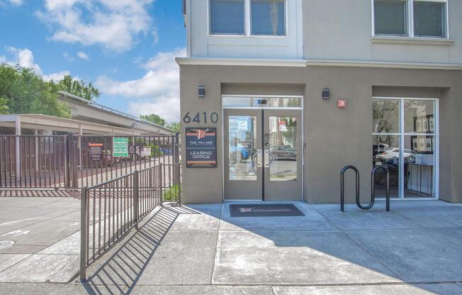 Leasing office entrance of The Villages, located at 6410, featuring large glass doors, a metal railing, and a bike rack in front. The building has a modern facade with a sign displaying the leasing office information, set against a blue sky with scattered clouds.