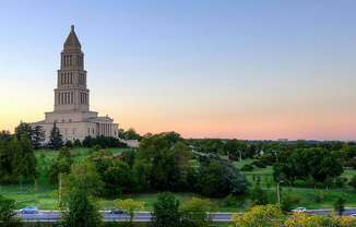 A large, towering building with a spire is surrounded by trees and greenery.