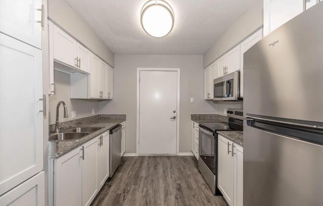 Modern kitchen with stainless steel appliances including a microwave, oven, and refrigerator. Features white cabinetry, a double sink, and gray countertops. The space has a neutral color palette and a bright overhead light, creating an open and inviting atmosphere.
