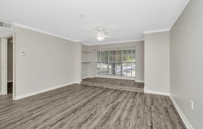 Spacious, empty living room with light-colored walls and a modern ceiling fan. Features large windows with blinds, allowing natural light to flow in. The floor is covered in laminate flooring, and there's a small step leading to a different level in the room. Minimalistic design with no furniture present.