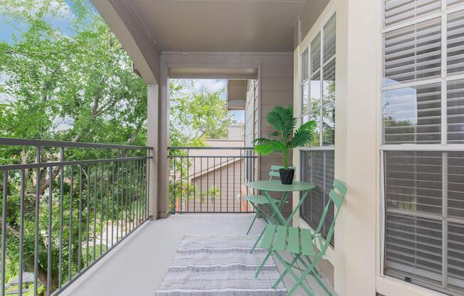 A sunny balcony with a small green table and two chairs, adorned with a potted plant. The space features a light-colored rug and has views of surrounding trees and buildings, creating a relaxing outdoor atmosphere.