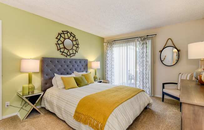 Model home bedroom with queen size bed, carpeting, nightstands and dresser with patio door on one side of room.