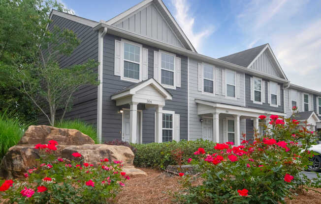 A house with grey siding and white trim is surrounded by red flowers and greenery.
