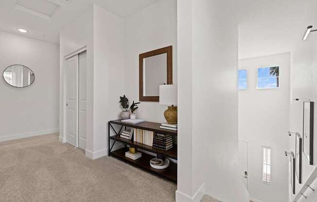 A hallway with a carpeted floor, a mirror, a framed picture, a console table with a vase and books, and a staircase.