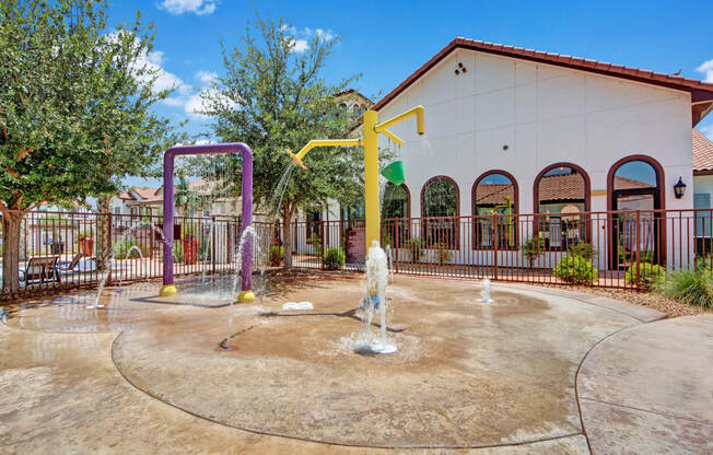 A fun outdoor splash pad here at Andalucia Villas with interactive water sprays and colorful play structures, surrounded by secure fencing, shade trees, and Spanish-style architecture that creates a safe and playful community space.