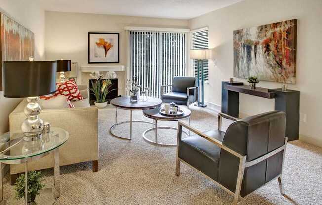 A welcoming living room here at Heritage Hills featuring neutral carpeting, a comfortable sofa with patterned accent pillows, and two modern armchairs arranged around nested round coffee tables. The space includes a fireplace with framed artwork above, large windows and a sliding glass door with vertical blinds, a console table with abstract art, and soft ambient lighting throughout.
