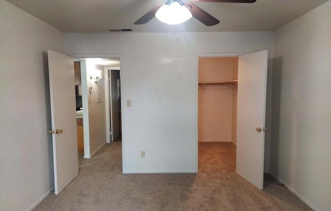 A spacious room featuring light-colored walls and a ceiling fan. Two white doors are visible, one leading to a closet and the other to a hallway. The floor is covered in beige carpeting, creating a cozy atmosphere. The room is well-lit with natural light.