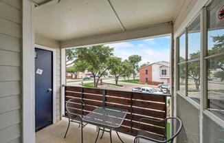 A small balcony with a table and two chairs overlooks a green area with trees. A blue door is visible to the left, while a view of nearby buildings and parked cars can be seen in the background against a partly cloudy sky.