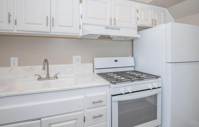 A modern kitchen featuring white cabinets, a stainless steel sink, a gas stove with an oven below, and a white refrigerator. The countertops are light-colored with a marble-like pattern, and the wall is painted a soft beige.