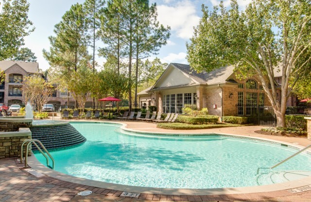 View of Fenced-In Pool Area with Loungers, Landscaping, and View of Apartment Buildings in Background at Raveneaux Apartments