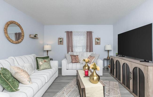 Cozy living room featuring a light-colored sofa and armchair with decorative pillows. A coffee table with vases of flowers sits in the center. The room has a large TV on a wood cabinet, two lamps, a round mirror, and light curtains, creating a warm and inviting atmosphere.