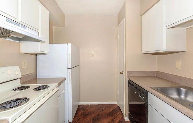 A small kitchen featuring white cabinets, a white refrigerator, a white oven with a stovetop, a black dishwasher, and a stainless steel sink. The walls are painted in a light beige color, and the floor is covered with dark wood laminate. The space is compact and well-organized.