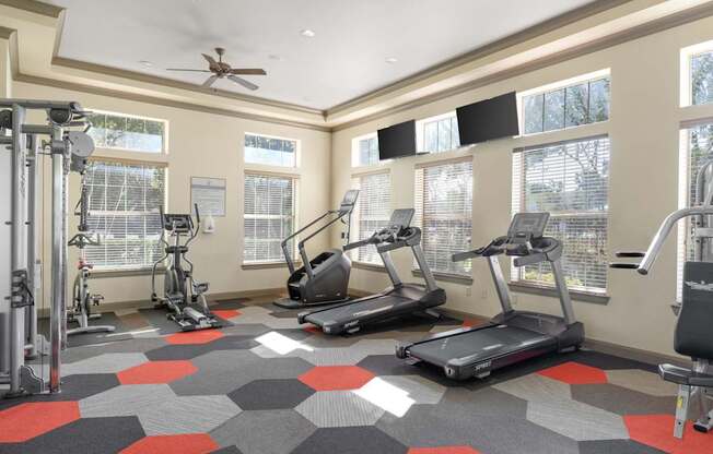 A fitness room here at Landing at Mansfield with treadmills, ellipticals, weight machines, and gray-and-red hex tile flooring, plus large bright windows and wall-mounted TVs.