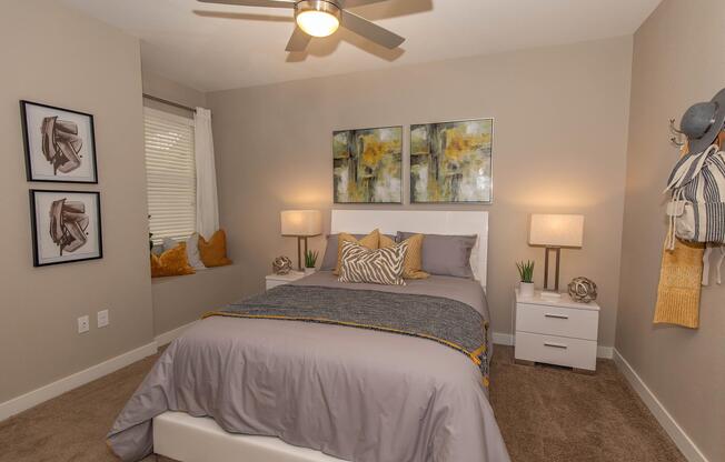 A cozy, modern bedroom featuring a queen-sized bed with gray bedding and yellow accents. Two decorative lamps sit on bedside tables next to the bed, which is adorned with patterned pillows. Abstract art hangs above the headboard. A window with light curtains and a small nook with cushions add charm to the space.