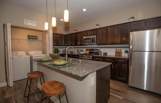 Modern kitchen featuring dark wood cabinetry, stainless steel appliances, and a granite countertop island with two wooden stools. A laundry area is visible to the left, and decorative elements, including plants and a fruit bowl, add a touch of warmth to the space. Bright pendant lights hang above the island.