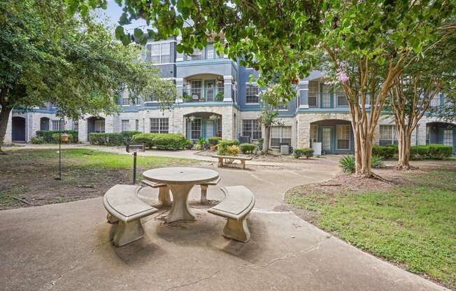 A concrete table and chairs are in the foreground of a courtyard with a building in the background.