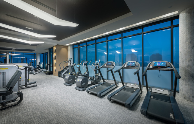 Bank of treadmills, ellipticals, and stair climbers in front of a large window wall with a city skyline view.
