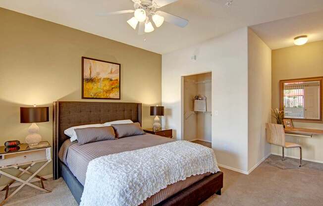 A bright bedroom here at Dobson Towne Center featuring plush carpet, a neutral accent wall, and a ceiling fan with light overhead. A stylish upholstered bed with textured bedding is framed by matching nightstands and lamps, while an open closet and built-in vanity area with mirror create a functional and comfortable retreat.