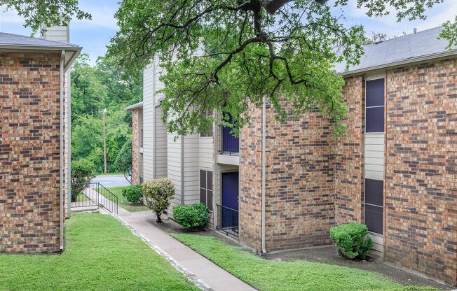 A view of a brick apartment community surrounded by greenery, featuring a pathway leading towards the entrance of the buildings, with a tree overhead and a glimpse of a pool in the background.