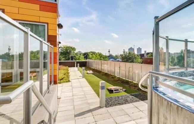 A sunlit rooftop walkway here at Blue and Lime featuring a paved path alongside glass railings, landscaped greenery, and an open-air seating area. The space overlooks surrounding treetops and city views, with modern building details and clean lines that create a relaxed outdoor setting for residents to enjoy fresh air and skyline scenery.