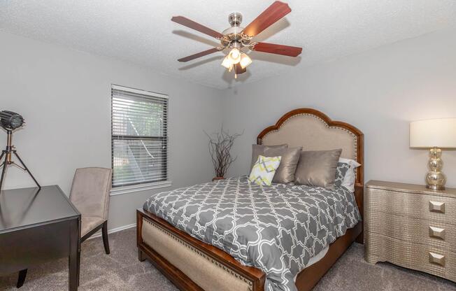 Cozy bedroom featuring a comfortable bed with decorative pillows, a stylish headboard, and a nightstand with a lamp. A sleek desk and a chair are positioned nearby, and a ceiling fan adds to the room's ambiance. Natural light enters through a window with blinds, creating a warm and inviting atmosphere.