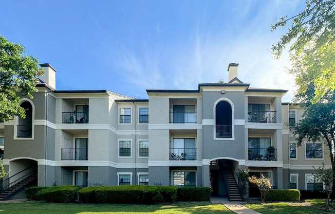 A light gray apartment building with balconies at Saxony at Chase Oaks Apartments in Dallas, TX, surrounded by manicured lawns and trees under a clear blue sky.