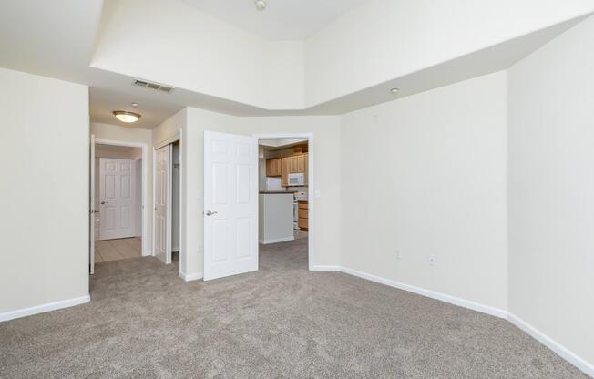 A spacious, well-lit room with beige walls and plush carpet. The image shows an open area leading to two doors on the left and an entryway to a kitchen in the background. The ceiling is slightly sloped, adding character to the space. The overall feel is inviting and ready for furniture.