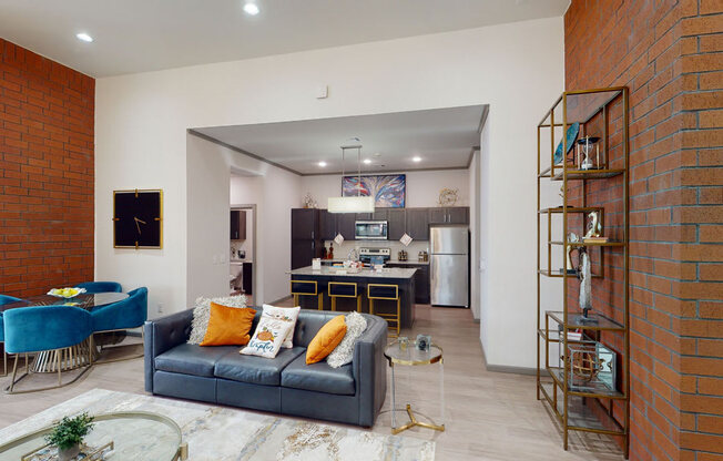 Contemporary resident lounge with a blue couch, orange and white pillows, and a glass table. A modern kitchen with dark cabinets is visible in the background.