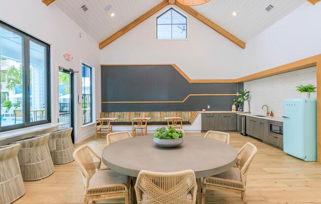 Modern interior of a communal space with high ceilings and wood accents. A large round table with a gray surface and woven chairs is centered in the room. To one side, there's a kitchen area featuring a retro-style refrigerator, cabinetry, and a plant decoration. Large windows provide natural light.