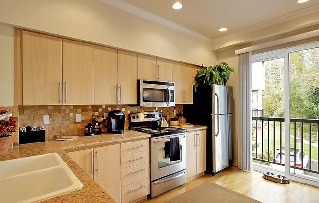 2-Bedroom Apartments in Kenmore, WA - Northshore Townhomes - Kitchen with Hardwood Flooring, Stainless Steel Appliances, Tan Wood-Tone Cabinetry, Slab Granite Countertops with Tile Backsplash, Decorative Plants, and Sliding Glass Door with Balcony Access.